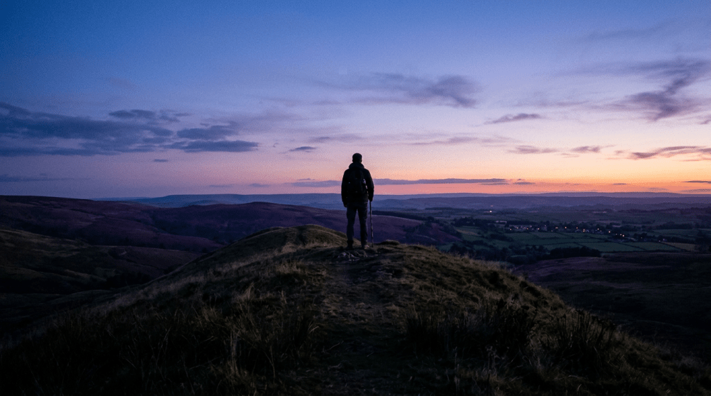 Person standing on a grassy hilltop looking at sunset over rolling hills and a distant village